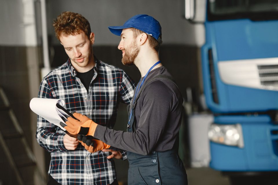 A mechanic in a blue cap walks a customer through paperwork on a clipboard in the shop
