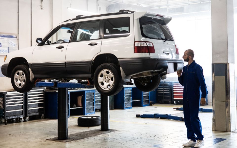 A mechanic in blue coveralls looks up at a white SUV raised on a lift in a bright, tidy repair bay