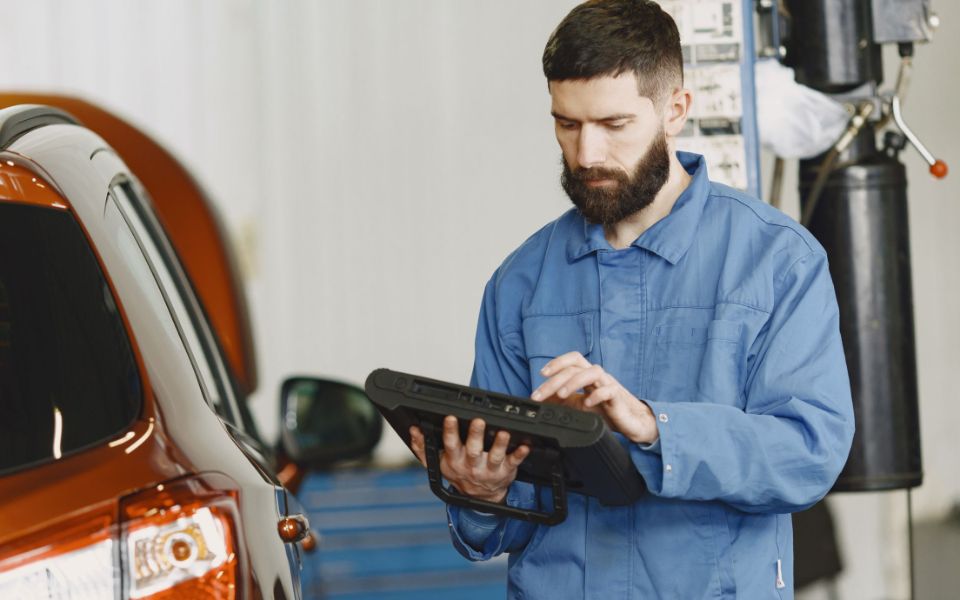 A bearded mechanic in blue coveralls taps through a tablet beside an orange SUV in the shop