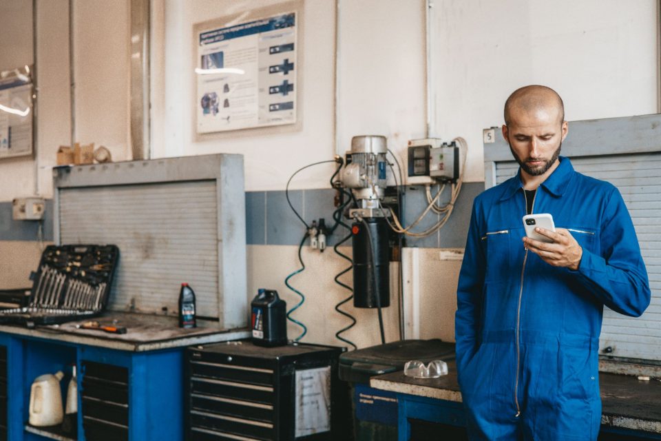 A mechanic in blue coveralls checks his phone beside a workbench and tool chest in the shop