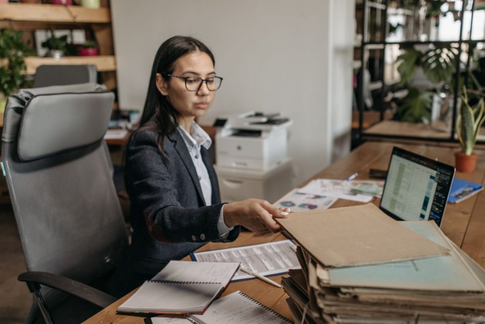 A business owner in glasses works through a stack of file folders beside a laptop showing a spreadsheet