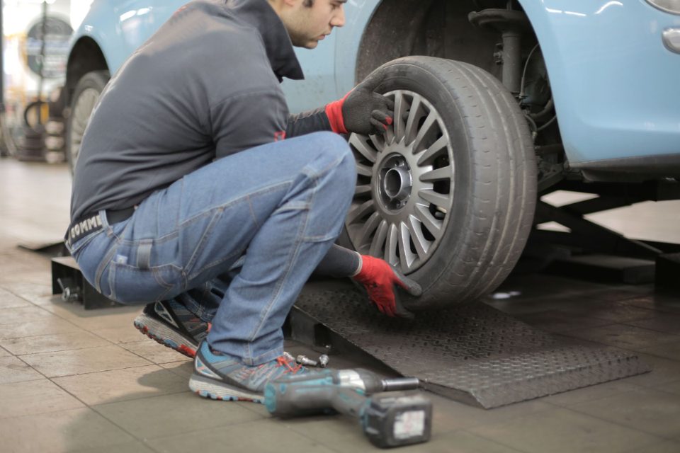 A technician in work gloves crouches beside a light blue car on a lift and steadies an alloy wheel on the hub